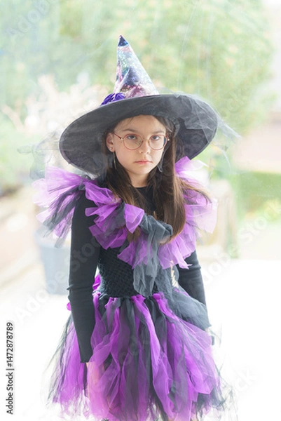 Obraz Cute girl in a witch costume and glasses poses standing in front of a large bright panoramic window. Concept of a children's party and childhood