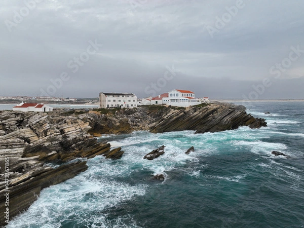 Fototapeta Low drone view of buildings on edge of rugged rocky cliffs with crashing waves
