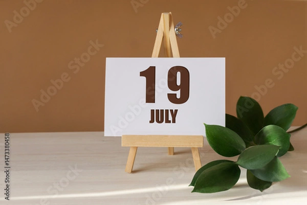 Fototapeta July 19th. Day 19 of month, Calendar date. Green branch, easel with the date and month on desktop. Close-up, brown background. Summer month, day of year concept