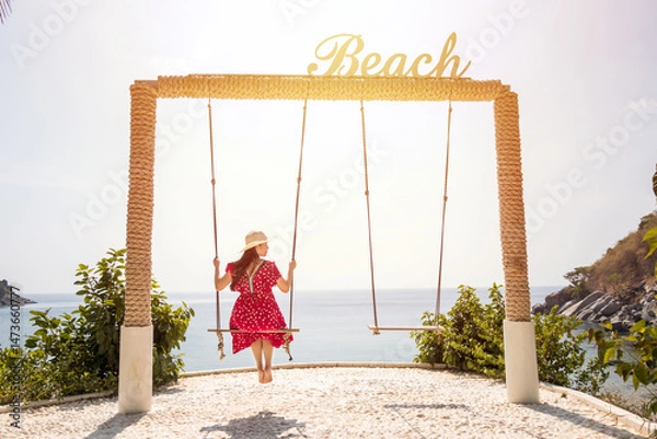 Fototapeta back view of Asian woman in red maxi dress sitting on a swing with a sign "Beach" on top, sea view at the background. happy summer travel concept
