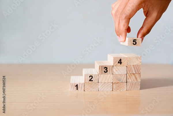 Obraz Close-up of a woman's hand placing the fifth wooden block on a staircase-like stack on a wooden table, symbolizing growth, progress, or step-by-step success