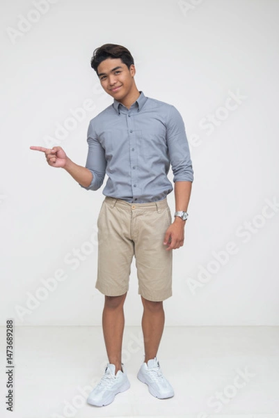 Fototapeta Southeast Asian man in casual clothes smiles while pointing to the left, appearing friendly and playful. Isolated on a light background.
