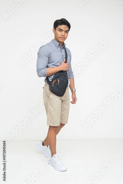 Fototapeta Southeast Asian man in smart casual outfit walks confidently while holding a black chest bag across his body. Isolated on a light background.