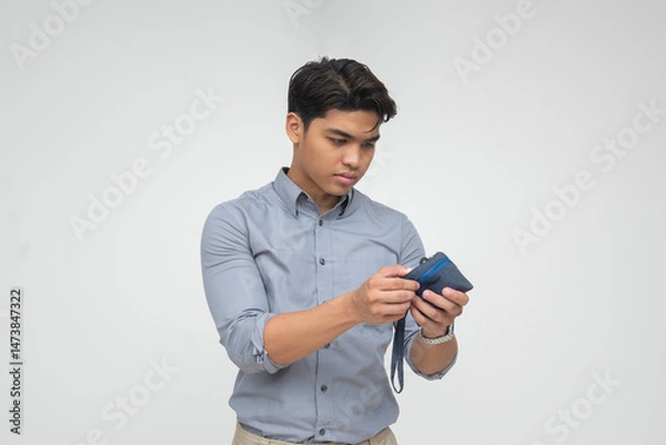 Fototapeta Southeast Asian man closely inspects a blue coin purse, appearing to look for any remaining change inside. Isolated on a light background.