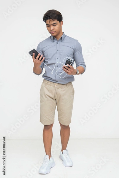 Fototapeta Southeast Asian man looks at his smartphone connected to a power bank with cable while standing, suggesting charging on the go. Isolated on a light background.