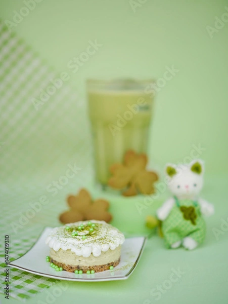 Fototapeta Matcha menu. Matcha greentea cheesecake mini tart on the foreground and Matcha green tea latte on the background. Soft focus on the cheesecake. Shamrock cookies decorations for Saint Patrick's day