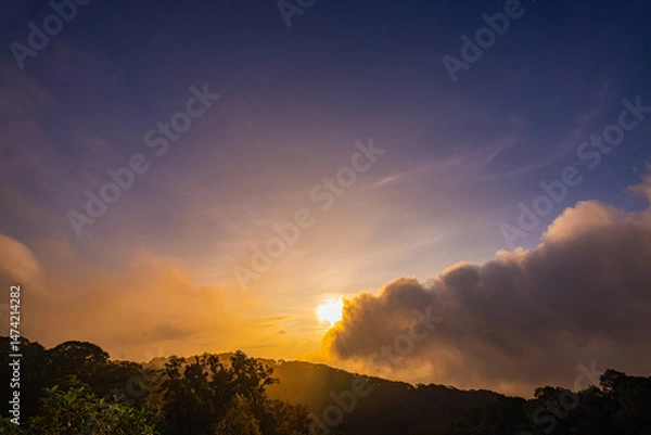 Obraz stunning mountainous landscape at sunrise with vibrant orange and pink hues illuminating the clouds against a blue sky. layer of mist settled in the valley, giving the scene a dreamy.