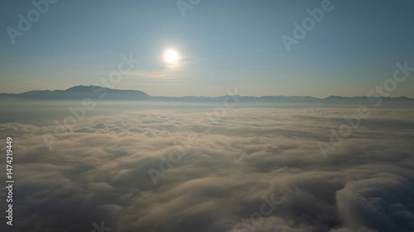 Fototapeta A serene view of the early morning sun rising above a vast sea of clouds, with distant mountain peaks faintly visible on the horizon, creating a peaceful and ethereal landscape. Flying over the cloud