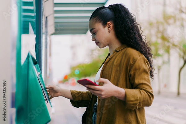 Fototapeta Focused Latina woman using self-service kiosk at transit station while holding a smartphone. Female tourist represents modern transportation, digital solutions, and convenience in everyday city life