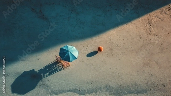 Fototapeta A beach umbrella casts a long shadow on the sand, accompanied by a chair and an orange ball nearby. This tranquil coastal scene captures a moment of relaxation under the sun