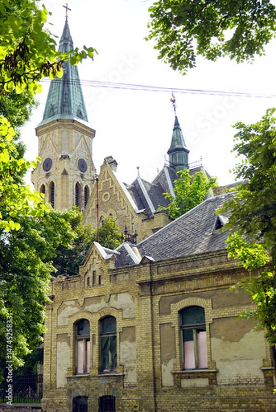 Fototapeta Beautiful old abandoned church in a European city: view of the brick facade of the building with neo-Gothic decoration surrounded by trees. Evangelical Church (Evangelička Hrišćanska Crkva) in Pancevo