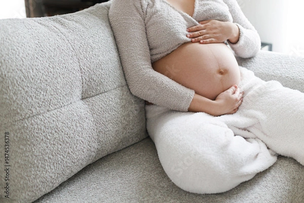 Fototapeta Pregnant woman in cozy clothes hugging baby bump and relaxing on grey couch in modern living room. Fertility and maternity. Happy Mothers day. Love and care concept