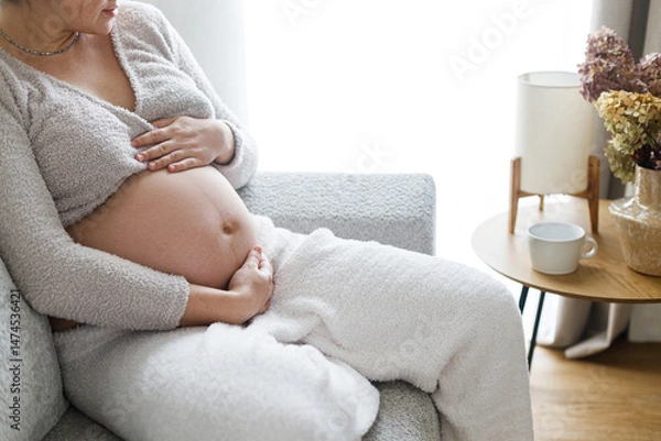 Fototapeta Pregnant woman hugging baby bump, sitting and relaxing on cozy couch in modern living room. Happy Mothers day. Love and care concept. Fertility and maternity