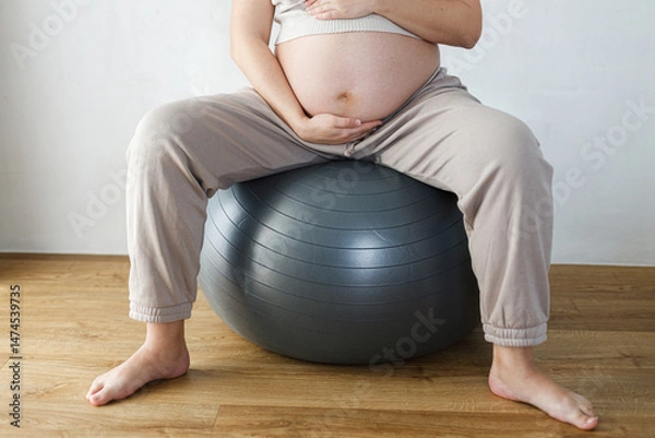 Fototapeta Pregnant woman holding baby bump and exercising on fitball at home. Pregnant yoga and wellness. Fertility and maternity. Pregnancy fitness.  Love and care concept