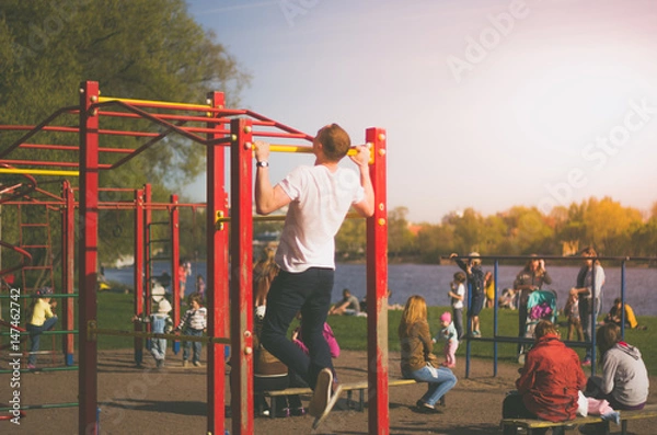 Fototapeta A man is practicing on a bar in a public park. A man pulls himself up on a bar in a street playground