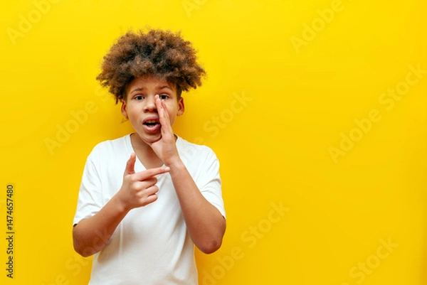 Fototapeta shocked curly african american boy whispering and announcing secret information on yellow isolated background, surprised teenager in white t-shirt telling secret and pointing at copy space