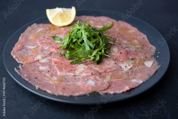 Fototapeta Marbled beef carpaccio with arugula, parmesan cheese, olive oil and lemon. A traditional classic appetizer made from thinly sliced raw meat. Close-up, selective focus, black background.