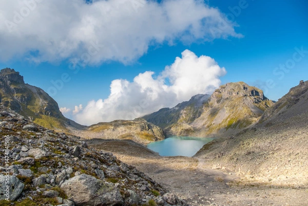 Fototapeta Captivating landscape of Pizol Wildsee lake in the Swiss Alps, featuring tranquil turquoise water amid rugged peaks enveloped by mist and clouds, creating a dramatic mountain scene