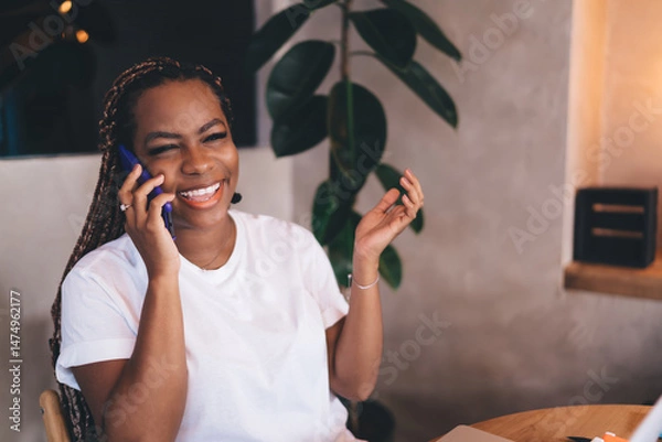 Fototapeta Close-up of cheerful young Black woman laughing during phone conversation, sitting in cozy workspace with relaxed attitude and tech gear in background