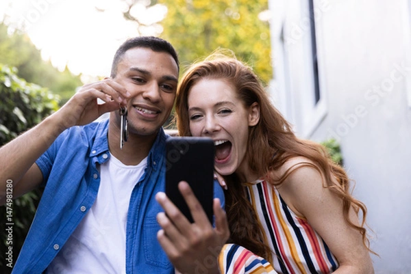 Obraz Young couple celebrating new home purchase, holding keys and taking selfie outside