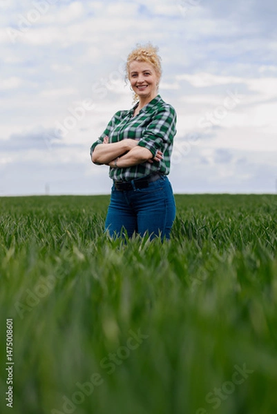 Obraz Adult female farmer standing in green wheat field looking at camera.