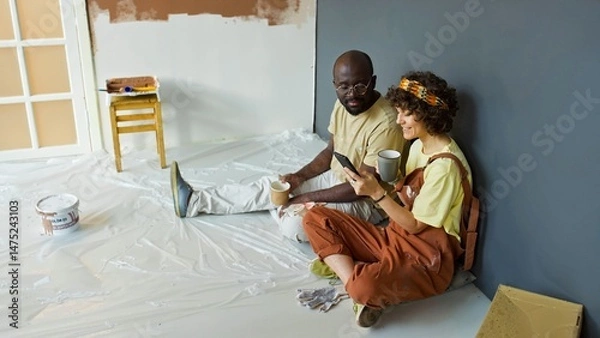 Fototapeta Black man and Hispanic woman enjoying coffee and looking at smartphone while taking break from painting in renovated room