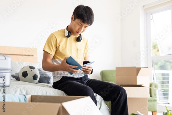 Obraz Young man reading book on bed surrounded by moving boxes and soccer ball