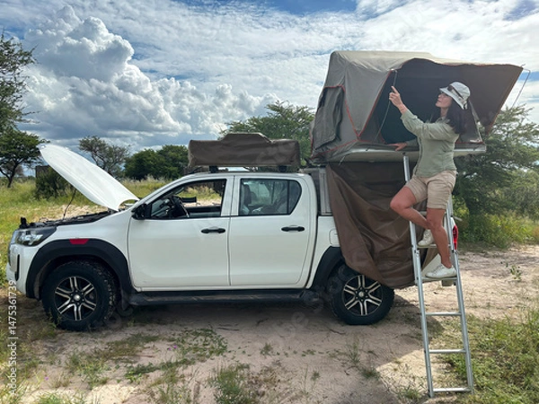 Fototapeta A dark-haired woman in safari attire stands on a ladder adjusting a roof tent on an off-road vehicle in the African wilderness. Adventure in the Central Kalahari, Botswana. 