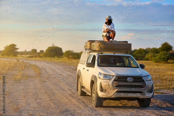 Fototapeta Woman on rooftop tent vehicle in a dry savannah, golden light, ready for wildlife photography. Clean composition with wide, open savannah and pastel sky.