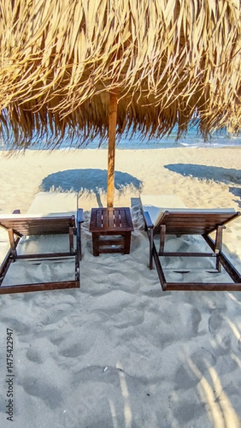 Fototapeta Mobile photo a calm and inviting beach scene with a thatched umbrella and lounge chairs on the sand. The gentle waves and clear sky create a serene and picturesque coastal environment.