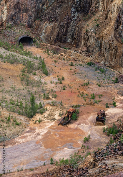 Fototapeta In the former copper mine of Falun Sweden, rusted vehicles lie amid a colorful red soil landscape of minerals. A bulldozer and crater showcase the site’s industrial past and nature's reclamation