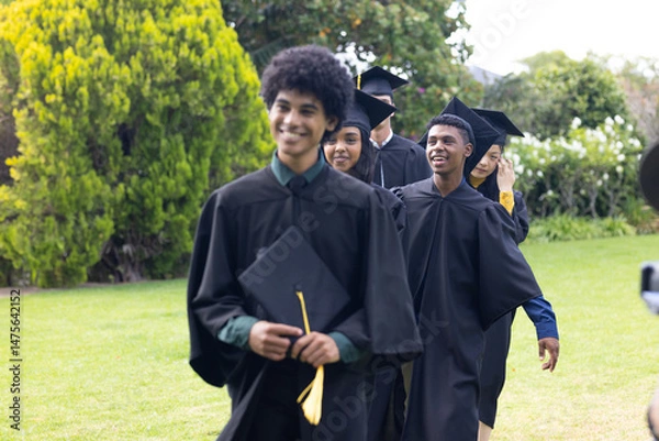 Obraz Graduating students in caps and gowns walking joyfully on green lawn