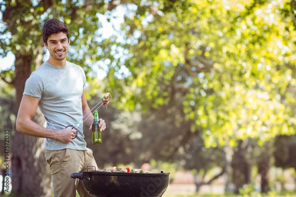 Obraz Charcoal barbecue grill cooking meat and vegetable skewers in sunlit park, with green bottle