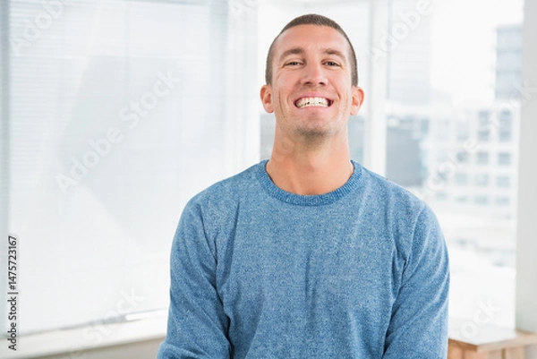 Obraz Smiling man wearing blue sweater standing in modern office studio, with blinds and city skyline