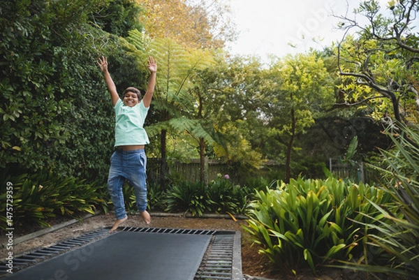 Fototapeta Boy jumping on in-ground trampoline in lush backyard garden, surrounded by ferns and shrubs