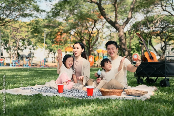 Fototapeta Happy family having a picnic and playing with bubbles together at the park