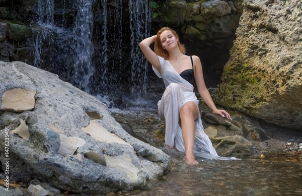 Fototapeta A girl in a transparent white dress sits on the rocks by the waterfall
