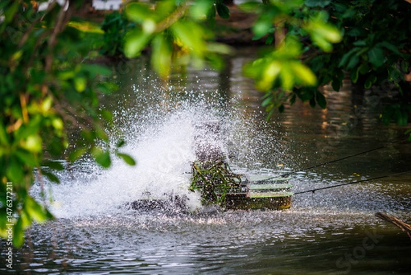 Fototapeta a water wheel aerator spinning and splashing in a pond, surrounded by green leaves and trees, creating oxygen for aquaculture and improving water quality in rural area