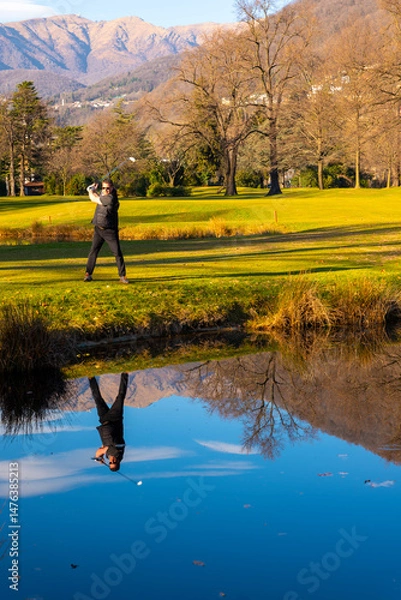 Fototapeta Male Golfer Reflected in a Water Pond and Hitting the Golf Ball on Fairway on Golf Course with Mountain in a Sunny Day in Switzerland.