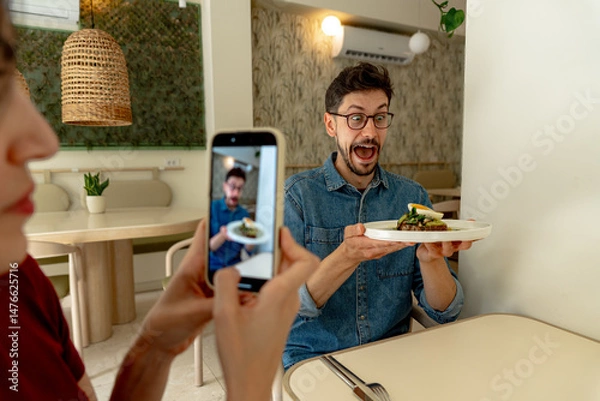 Fototapeta Man showing his gourmet dish with surprised expression while woman taking photo with phone in modern restaurant, sharing food experience