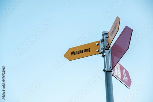 Fototapeta Signpost displaying directional arrows, including a yellow Wanderweg sign, set against a bright blue sky, representing outdoor adventure and navigation choices