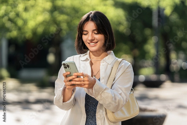 Fototapeta A summer day out. A smiling woman checking an app on her phone in the city. Browsing online shops, chatting with friends and family or checking the map for navigation