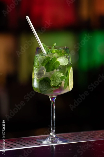 Fototapeta A refreshing green Mojito cocktail, filled with mint leaves, lime slices, and ice, is served in a tall glass with a white straw.