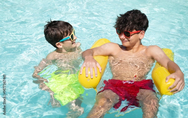 Fototapeta Two brothers wearing sunglasses are enjoying their summer holidays, playing in a swimming pool with an inflatable pool mattress
