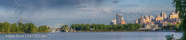 Fototapeta Modern residential buildings and pedestrian bridge on the Dnipro River in Kyiv at sunset. Peaceful cityscape with calm water, trees, and dramatic sky.