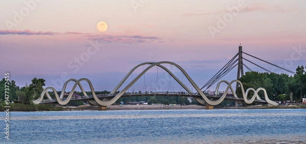 Fototapeta A glowing full moon ascends behind Kyiv’s elegant pedestrian bridge, casting golden reflections across the calm Dnipro River in a serene embrace of night and light.