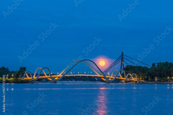 Fototapeta Full moon rises over the Dnipro River in Kyiv, glowing behind a modern pedestrian bridge. City lights and calm water create a peaceful and dreamy evening atmosphere.