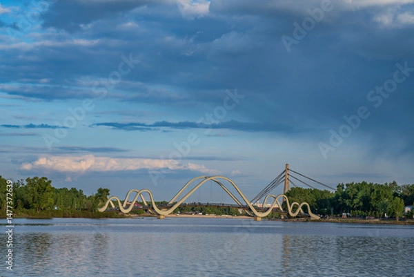Fototapeta Dramatic clouds hover over the pedestrian bridge on the Dnipro River in Kyiv at sunset. Tranquil cityscape with calm water and soft golden light.