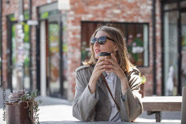 Obraz Beautiful woman in sunglasses holding disposable cup of coffee while sitting on open veranda in cafe