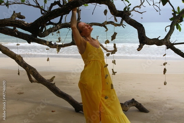 Obraz An adult beautiful blonde woman in a yellow long dress walks on the shore of the Andaman Sea and poses under a tree decorated with seashells.